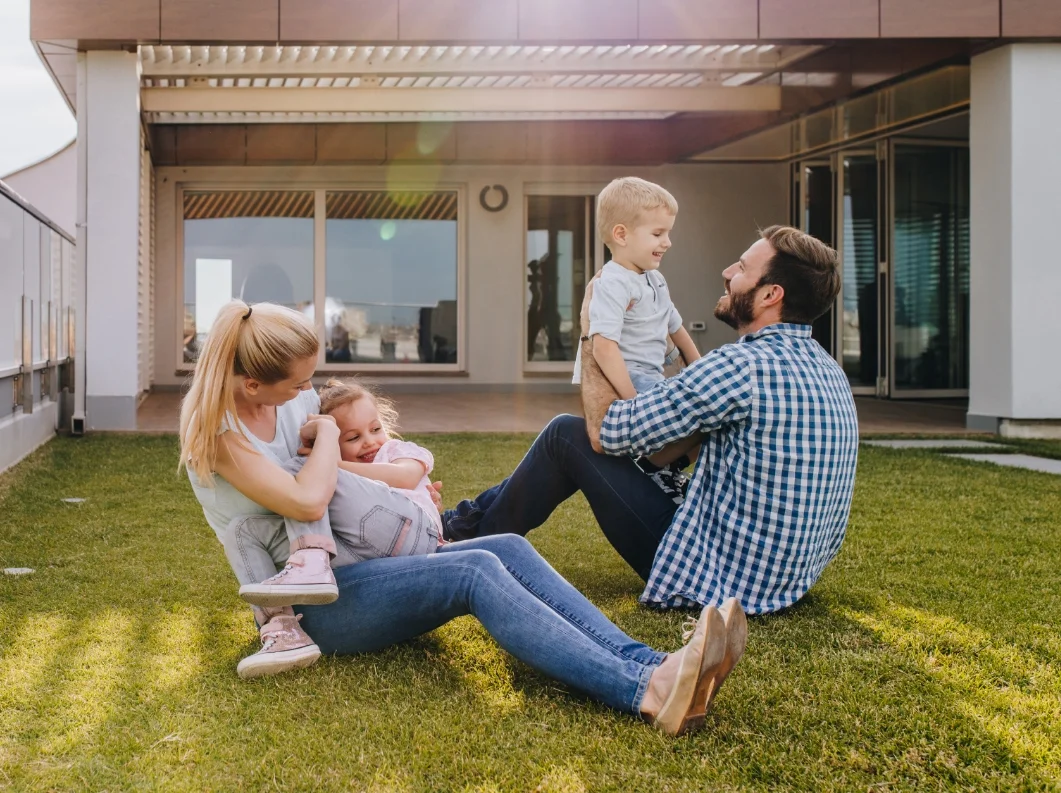 Eine vierköpfige Familie spielt lachend auf einer Rasenfläche vor einem modernen Wohnhaus.