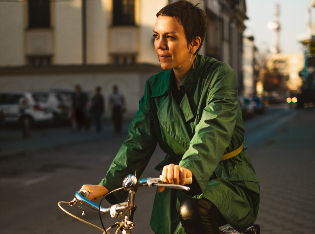 Frau in grünem Mantel fährt bei tiefstehender Sonne mit einem Fahrrad durch die Stadt.